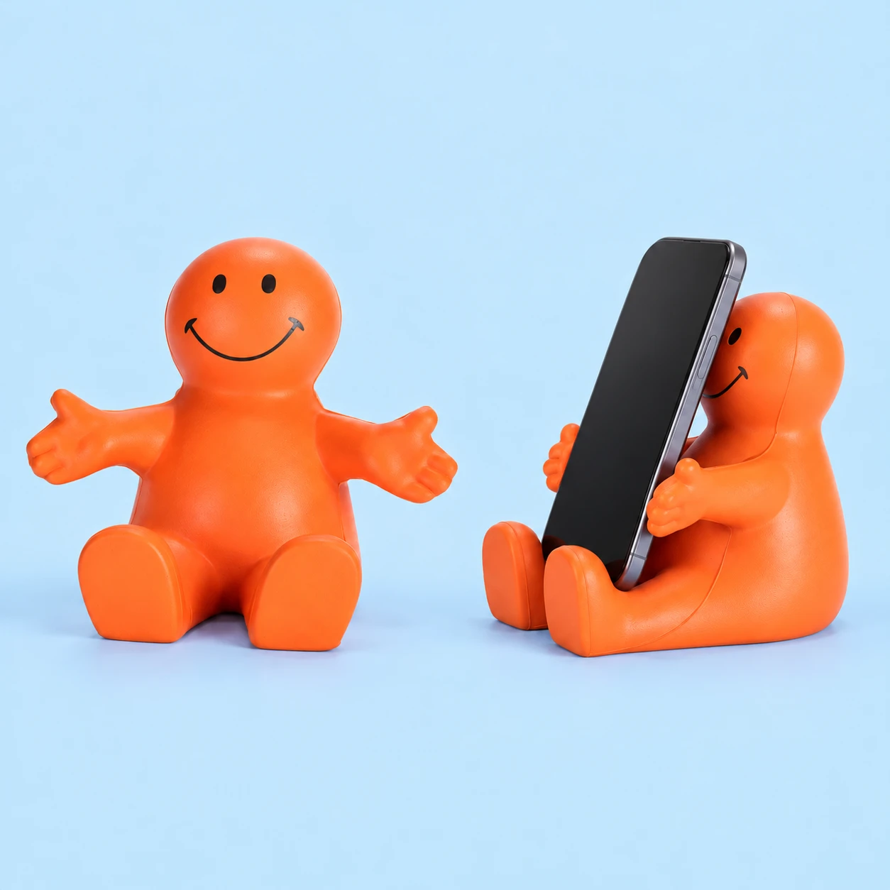 Orange smiling seated foam phone cradle, shown complete at left and in an illustrative portrait-phone resting arrangement at right; phone not included, square composition