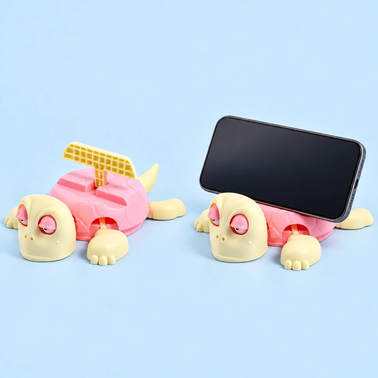 Pink-shell turtle phone stand with folding T-shaped backrest, shown alone and supporting a landscape phone, square composition