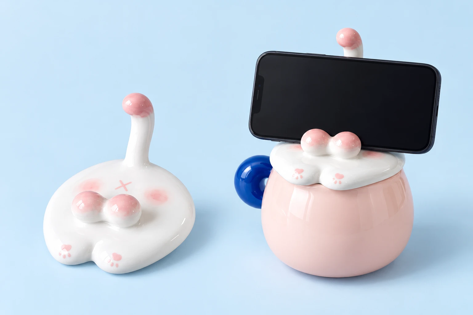 Pink cat-tail ceramic mug lid on pale blue, shown empty at left and resting across a compatible dry pink mug rim while supporting a landscape phone at right. Mug and phone are demonstration props and are not included.