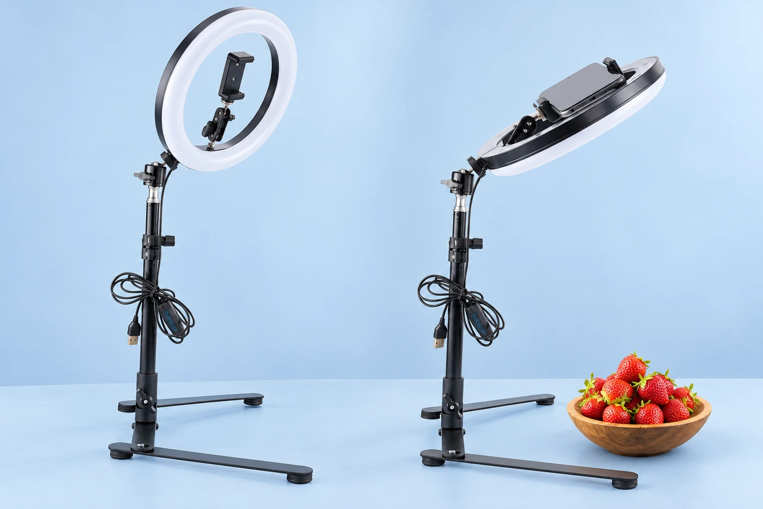 Complete black twin-runner overhead phone and 10-inch ring-light kit, empty at left and holding a demonstration phone above strawberries at right on pale blue. Both lights are off and cables unplugged. Phone and props not included.