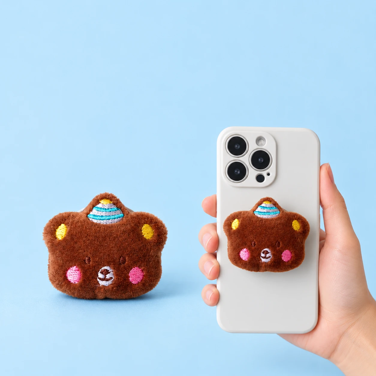 Brown plush bear-head phone grip with embroidered party hat and closed phone-back placement, square composition
