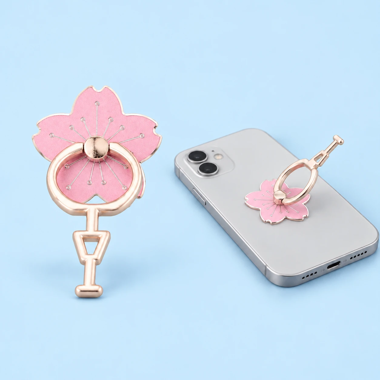 Light-pink cherry-blossom phone ring with its long continuous foot, complete and mounted on a flat-lying phone in a same-model use illustration, square composition