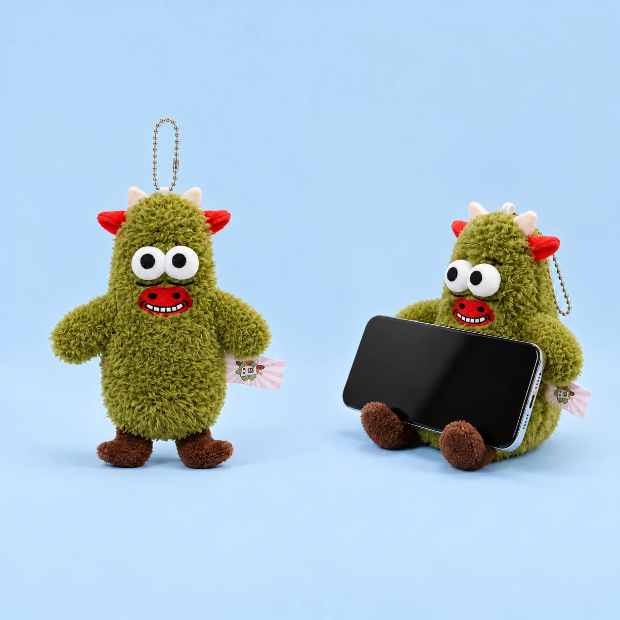 Green horned cow plush phone rest with red ears and muzzle, shown complete and supporting an illustrative landscape phone, square composition