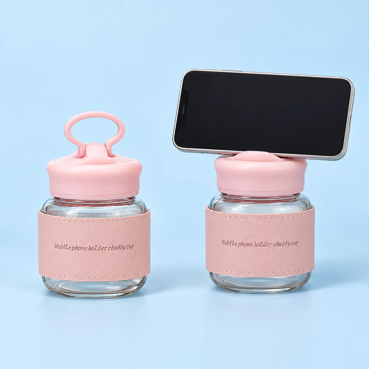 Two views of the same complete empty pink 300 mL glass cup with matching sleeve, raised phone-rest lid and oval carry loop: unoccupied on the left and supporting a landscape demonstration phone on a stationary pale-blue tabletop on the right. One cup is offered; phone not included.