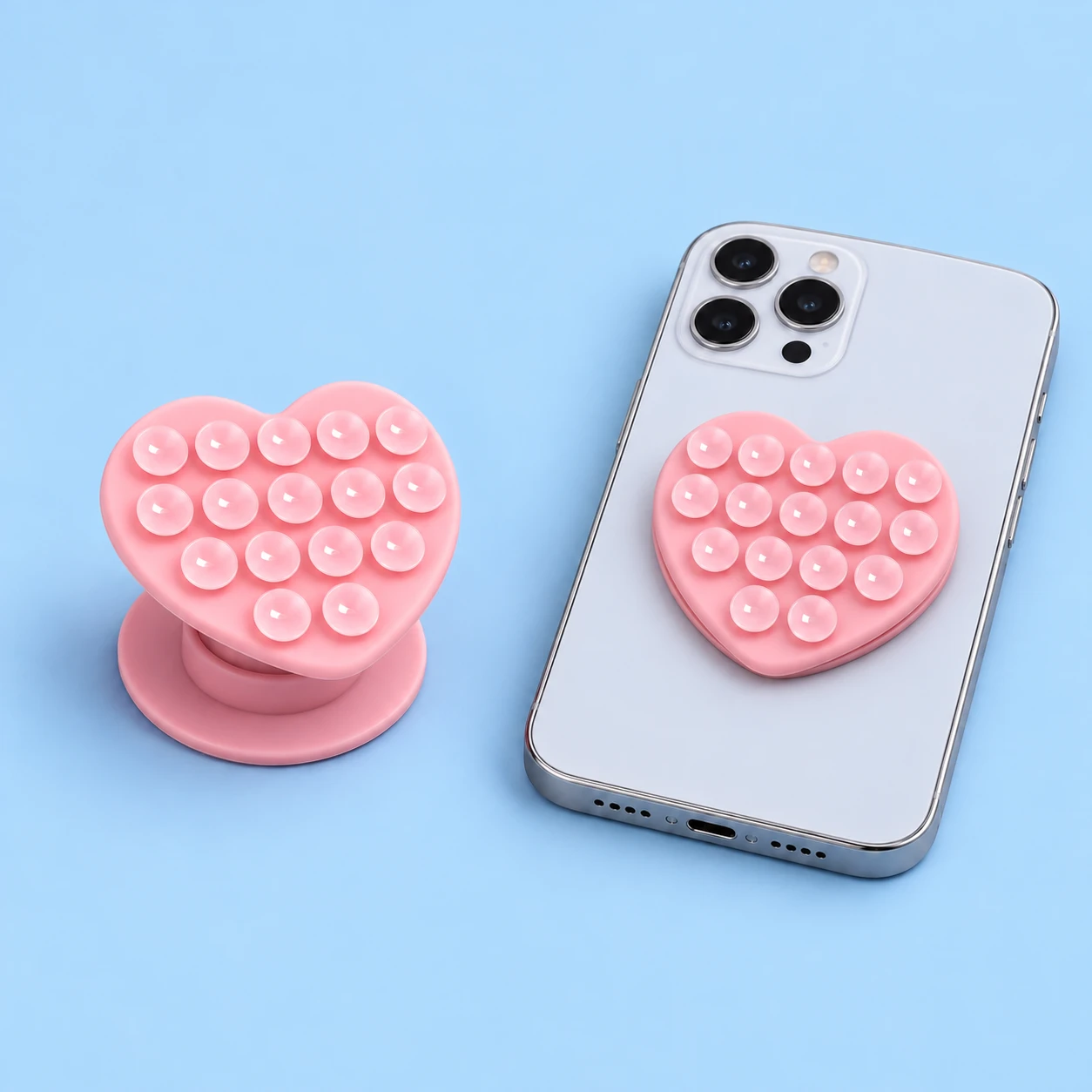 Pastel pink heart-shaped expandable phone grip with sixteen suction cups and round base, shown alone and closed on a phone, square composition