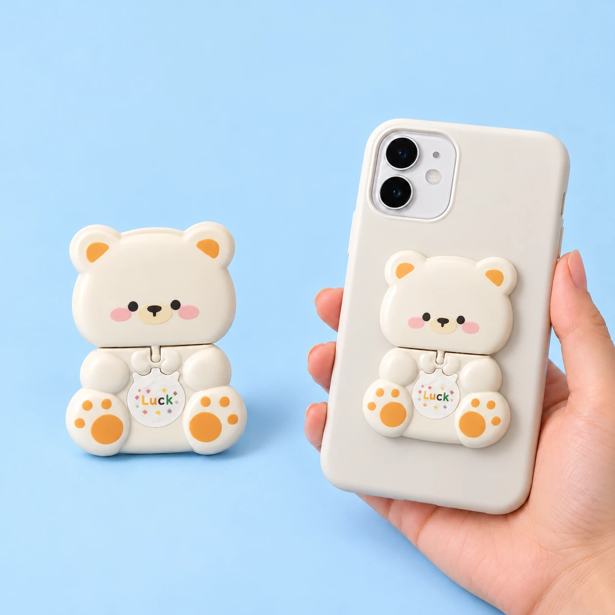 Cream bear-shaped folding mirror phone kickstand with orange paw details, shown closed alone and on a phone case, square composition