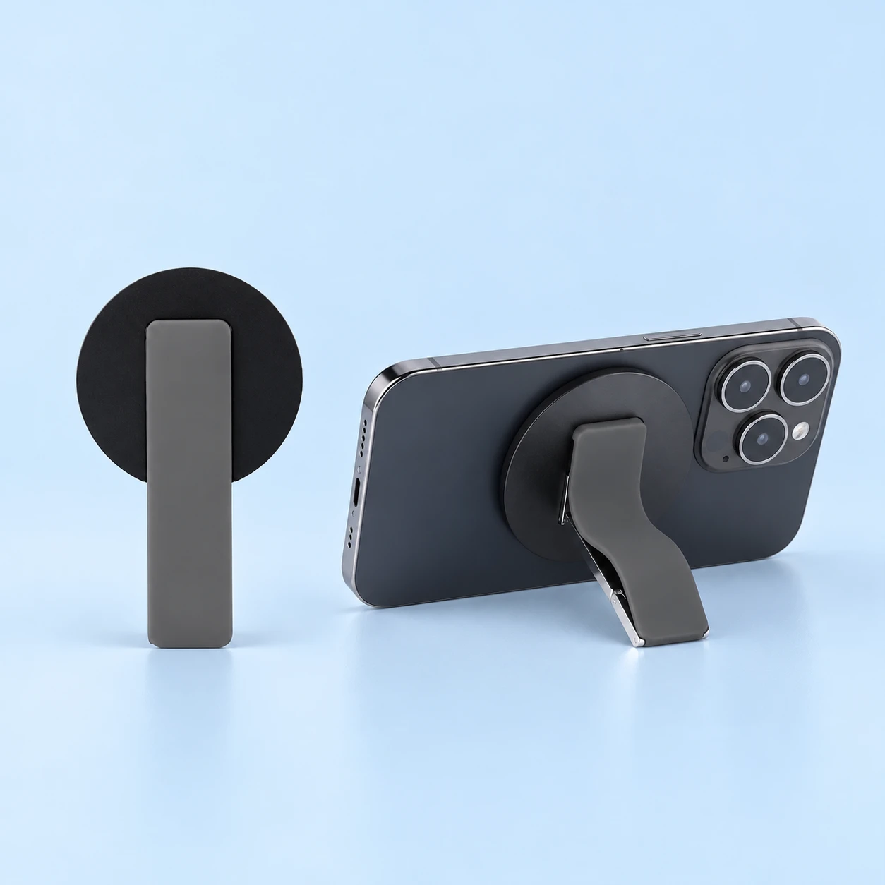 Gray magnetic folding finger band shown folded on the left and supporting a landscape phone from behind on the right, on pale blue., square composition